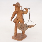 Arte popular brasileira. Escultura em barro representando pescador ambulante, trajando chapéu de abas largas e bolsa a tiracolo, portando balança suspensa por fios metálicos com peixes modelados sobre os pratos, assente sobre base quadrangular. Assinada João Ezequiel. Medidas: Alt.: 21 x Comp.: 14 x Larg.: 9 cm.