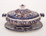 Terrina com tampa e travessa em faiança esmaltada, decoração em azul cobalto e manganês sobre fundo branco, apresentando reservas com motivos vegetalistas, aves e arquitetura estilizada, inspiradas na cerâmica hispano-mourisca. Corpo oval com alças laterais integradas e tampa com pomo moldado. Acompanha travessa de apresentação. Marca da manufatura na base. Medidas: Alt.: 12,5 x Comp.: 23 x Larg.: 14,5 cm.