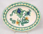 Travessa oval em faiança esmaltada, decoração policroma em tons de verde, azul e amarelo, apresentando composição central de flores estilizadas e volutas vegetalistas, com larga cercadura ornamentada por motivos repetitivos semicirculares. Medidas: Comp.: 52 x Larg.: 41 cm.