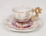Xícara miniaturizada com respectivo pires em porcelana, decoração em reserva branca com motivos florais em douração, faixa em tom vinho e aplicações douradas em relevo, alça vazada de desenho geométrico. Apresenta restauro na alça, desgastes na douradura e policromia. Medidas: Alt.: 3 x Comp.: 6 x Larg.: 4 cm. ; Medidas pires: Diam.: 8 cm.