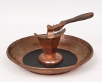 Quebra-nozes com bandeja, executado em cobre martelado, composto por bandeja circular com centro em material sintético na cor preta e suporte central integrado para quebra-nozes articulado com mandíbulas serrilhadas. Apresenta desgastes e sinais de uso. Medidas: Alt.: 10 x Diam.: 12 cm.