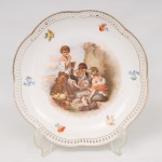 Prato decorativo em porcelana alemã Bavaria, borda recortada com vazados e contornada por filete dourado, decorado ao centro em policromia com cena inspirada na obra "Meninos Jogando Dados" de Bartolomé Esteban Murillo,  e reservas florais distribuídas pela aba. Marca da manufatura no verso. Medida: Diam.:  25 cm.
