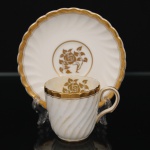 Minton, Staffordshire - Inglaterra. Excepcional xícara para café em porcelana inglesa da década de 1950, padrão Gold Rose. Esmaltagem vítrea de tom branco perolado, realçado por profusa ornamentação com faixas e filetes à ouro, fundo interno contendo flor em reserva. Bojo aproximadamente cilíndrico com alça lateral em voluta, ricamente modelado com frisos em espiral relevados na própria pasta; padrão decorativo replicado no pires. Marca da manufatura no verso. Medidas: Xícara: Altura:  x Comprimento:  x Largura:  cm; Pires: Diâmetro:  cm; Prato: Diâmetro: 17,5 cm. Apresenta marcas de uso e do tempo, vide foto.