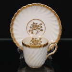Minton, Staffordshire - Inglaterra. Excepcional xícara para café em porcelana inglesa da década de 1950, padrão Gold Rose. Esmaltagem vítrea de tom branco perolado, realçado por profusa ornamentação com faixas e filetes à ouro, fundo interno contendo flor em reserva. Bojo aproximadamente cilíndrico com alça lateral em voluta, ricamente modelado com frisos em espiral relevados na própria pasta; padrão decorativo replicado no pires. Marca da manufatura no verso. Medidas: Xícara: Altura:  x Comprimento:  x Largura:  cm; Pires: Diâmetro:  cm; Prato: Diâmetro: 17,5 cm. Apresenta marcas de uso e do tempo, vide foto.