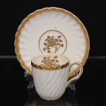 Minton, Staffordshire - Inglaterra. Excepcional xícara para café em porcelana inglesa da década de 1950, padrão Gold Rose. Esmaltagem vítrea de tom branco perolado, realçado por profusa ornamentação com faixas e filetes à ouro, fundo interno contendo flor em reserva. Bojo aproximadamente cilíndrico com alça lateral em voluta, ricamente modelado com frisos em espiral relevados na própria pasta; padrão decorativo replicado no pires. Marca da manufatura no verso. Medidas: Xícara: Altura:  x Comprimento:  x Largura:  cm; Pires: Diâmetro:  cm; Prato: Diâmetro: 17,5 cm. Apresenta marcas de uso e do tempo, vide foto.