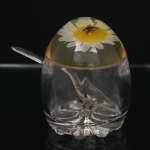 Açucareiro com colher em acrílico transparente moldado, composto por recipiente cilíndrico, tampa semiesférica decorada com margarida branca encapsulada e inseto em resina, acompanhado de colher em acrílico. Medidas: Alt.: 11 x Diam.: 9 cm. ; Medidas colher: Comp.: 12,5 cm.