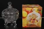 Bombonière com tampa, em vidro prensado incolor, apoiada sobre três pés integrados ao corpo, decorada em relevo com motivos vegetalistas e frutíferos. Acompanha caixa original de acondicionamento da marca Wincy, linha Classical. Medidas: Alt.: 20 x Diam.: 15 cm.