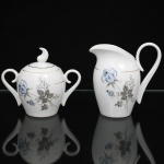 Porcelana Schmidt - Brasil(Fundada em 1945). Uma(01) leiteira e um(01) açucareiro em porcelana nacional, esmaltagem vítrea na cor branca, dotado de rica decoração com padrão de flores em estampagem, predominando as tonalidades azul, cinza e preto, realçado com frisos em douradura; bojo oval, asas laterais em voluta, bico vertedor revirado para fora, tampa ligeiramente abaulada encimada por pega com feitio de chama estilizada. Marca da manufatura ao fundo. Medidas: Leiteira Alt.: 14 x Comp.: 15 x Larg.: 10 cm; Açucareiro: Alt.: 14 x Comp.: 16,5 x Larg.: 10 cm.