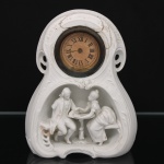 Relógio de mesa em porcelana de biscuit moldada em relevo, com caixa de formato rocaille, mostrador circular com numeração romana e composição escultórica representando casal em trajes do século XVIII disposto junto a mesa de apoio. Máquina mecânica com acesso posterior. Peça decorativa de inspiração romântica, produção europeia do final do século XIX ou início do século XX. Apresenta avarias, restauros e faltas, incluindo trincas na estrutura, perdas na porcelana, desgaste geral e mecanismo sem garantia de funcionamento. Condição conforme fotografias. Medidas: Alt.: 19 x Comp.: 15 x Larg.: 7 cm.