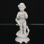 Escultura em porcelana de biscuit representando putto em pé junto a tronco estilizado, segurando ave nas mãos, sobre base moldada de contorno movimentado. Produção decorativa europeia, final do século XIX ou início do século XX. Apresenta marca em baixo-relevo na base. Estado de conservação com avarias, restauros e perdas, incluindo quebras e recomposições na região do pescoço, perdas na ave, desgastes e pequenas faltas na superfície. Condição conforme fotografias. Medidas: Alt.: 13,5 x Diam.: 6 cm.