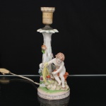 Abajur em porcelana policromada, representando figura infantil junto a tronco estilizado ornado por flores em relevo, servindo de haste para suporte de lâmpada. Base circular com friso de padrão grego. Marca na base Original Dresden, produção alemã do século XX. Apresenta instalação elétrica antiga. Condição conforme fotografias. Medidas: Alt.:  25,5 x Diam.: 10 cm.