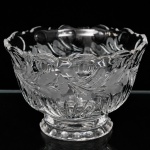 Fruteira em cristal incolor prensado, de bojo hemisférico assente sobre pé anelar moldado com sequência círculos em baixo relevo; borda recortada apresenta contorno ondulado, acompanhando a composição decorativa do recipiente. Decorado por lapidação geométrica em gomos radiantes na porção inferior, associada a faixa contínua gravada a ácido com motivos florais e folhagens estilizadas, intercaladas por reservas em forma de conchas, interior reproduz a estrutura facetada da lapidação. Medidas: Alt.: 14 x Diam.: 21 cm.