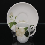 Xícara para café em porcelana, decoração transferida com rosas brancas e folhagens em tons de verde sobre fundo branco. Marca da manufatura no verso. Medidas: Xícara: Alt.: 6 x Comp.: 7,5 x Larg.: 5 cm. ; Pires: Diam.: 11,5 cm.