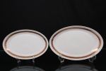 Porcelana, Steatita e Magnésia S/A, Campo Largo - PR(Fundada em 1954). Duas(02) travessas ovais em porcelana nacional da década de 1960/70; esmaltagem vítrea na cor branca, orla decorada por faixa de tonalidade bordô, ornamentada por arabescos fitomorfos dourados em repetição, cercado por frisos em douradura. Marca da manufatura no verso. Medidas: Comp.: 29 x Larg.: 20 cm. ;  Comp.: 33 x Larg.: 21 cm.