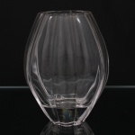 Nachtmann ou FX Nachtmann Bleikristallwerke, Oberpfalz - Alemanha(Fundada em 1834 por Michael Nachtmann). Vaso em cristal incolor moldado e lapidado em gomos verticais, corpo bojudo com borda recortada e base espessa facetada; desenho de linhas sóbrias e volumetria geométrica associado ao gosto moderno de influência Art Déco tardia e design escandinavo do pós-guerra, provavelmente produzido entre as décadas de 1940 e 1960. Marca da manufatura ao fundo. Medidas: Alt.: 20 x Comp.: 15 x Larg.: 10 cm.