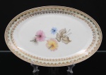 Porcelana Real, Mauá - SP(Fundada em 1943 por Fritz Erwin Schmidt). Travessa oval em porcelana nacional da década de 1950/60; esmaltagem vítrea na cor branca, orla circundada por faixa de acabamento nacarado, sobreposta por barrado contínuo de leques estilizados, guirlandas e frisos em douradura, centro dotado de bela composição botânica em estampagem policromada, predominando as tonalidades preta, rosa, azul e amarelo. Marca manufatura no verso. Medidas: Comp.: 28,5 x Larg.: 18 cm.