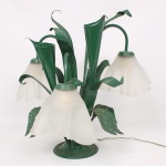 Luminária de mesa em metal com pátina de tonalidade verde, modelada na forma de lírios ou copos-de-leite estilizados, com três braços de luz e cúpulas em vidro fosco moldado. Corpo composto por folhas e flores em chapa metálica conformada, base circular e instalação elétrica interna. Parte elétrica não testada. Medidas: Alt.: 46,5 x Comp.: 48 cm.