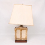 Abajur de mesa composto por base em cerâmica modelada com decoração simulando colmos de bambu, acabamento em tons marfim e dourado, apoiada sobre plinto de madeira. Cúpula trapeziforme revestida em tecido. Apresenta desgastes, manchas de uso e pequenas deformações na cúpula. Funcionamento não testado. Medidas: Alt. 62 x Comp.: 37 x Larg.: 26 cm.