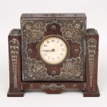 Relógio de mesa em madeira, com caixa arquitetônica de composição geométrica, apresentando mostrador circular com numeração arábica e moldura metálica. Corpo ornamentado por aplicações em relevo de temática floral e fitomórfica, distribuídas na face frontal e montantes laterais, executadas em metal. Estrutura apoiada sobre base escalonada, contendo mecanismo mecânico alojado no interior da caixa, acessível pela tampa posterior. Não está funcionando, apresenta desgastes e marcas de uso, vide foto. Medidas: Alt. 15,5 x Comp.: 18 x Larg.: 5 cm.