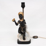 Abajur de mesa em porcelana moldada e policromada, representando figura feminina africana em posição sentada sobre formação rocaille. A personagem apresenta torso desnudo, adereços compostos por turbante, braceletes, argolas auriculares e colar, com aplicação de douração nos elementos ornamentais. Sustenta recipiente em uma das mãos, enquanto a estrutura elétrica emerge por haste metálica posterior destinada ao suporte da cúpula. Base de contorno recortado em material sintético, integrando o conjunto. Parte elétrica não testada, apresenta quebradura, demanda restauros. Medidas: Alt.: 36,5 x Comp.: 14 x Larg.: 11,5 cm.