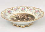 Belíssima fruteira para centro de mesa em porcelana, de formato circular recortado, assente sobre pé anelar vazado; orla decorada com esmaltação policroma com reservas florais entre volutas douradas, no medalhão central, cena representando a obra "Crianças Comendo Uvas e Melão"  de Bartolomé Esteban Murillo, bordas recortadas e contornadas com generosos realces a ouro. Medidas: Alt.: 8,5 x Diam.: 31,5 cm.