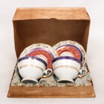 Duas(02) xícaras para chá com respectivos pires, em porcelana Schmidt, Santa Catarina, acondicionado em estojo. Corpos assentados sobre pé anelar, decorados com faixas em púrpura e azul-cobalto, acabamento perolizado e ornamentação em douração de motivos fitomorfos e rendilhados. Marca de manufatura na base. Medidas Xícara: Alt.: x Comp.: x Larg.: ; Pires: Diam.: