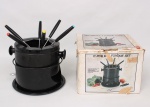 Aparelho para fondue composto por panela em metal esmaltado, suporte cilíndrico, queimador metálico com tampa reguladora e seis garfos de haste metálica com cabos em material sintético identificados por marcadores cromáticos. Acompanha caixa de acondicionamento original.