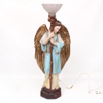 Tocheiro representando anjo, executado em estuque decorado com policromia e douração. Figura em posição frontal, trajando túnica em tonalidade azul com vestes inferiores em ocre, apoiada sobre base facetada, sustentando coluna central que serve de haste para suporte de iluminação. Asas posteriores amplas com generoso acabamento em douração. Cúpula superior em vidro prensado, moldado com decoração estriada radial e borda ondulada, encaixe metálico. Instalação elétrica posterior com interruptor no fio. Apresenta pequena quebradura na asa. Medidas: Alt.: 82 x Comp.: 17 x Larg.: 21 cm.