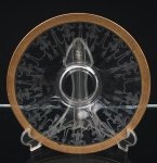 Prato para centro de mesa em cristal incolor moldado e gravado ao ácido, com cavidade central circular elevada e composição radial estilizada. Aba decorada por friso contínuo com motivos fitomórficos pendentes e volutas, executados por gravação fosca. Borda guarnecida por faixa em douradura, ornamentada com entrelaçamentos e elementos vegetalistas. Trabalho de estética Art Déco, primeira metade do século XX. Medida: Diam.: 27 cm.