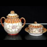 Um(01) açucareiro e uma(01) manteigueira em porcelana japonesa, bojo oval assente sobre base circular, decoração policromada e em relevo composta por reservas florais estilizadas sobre fundo perolizado, faixa superior ornamentada por filetes verticais em laranja, azul, verde, branco e dourado, tampa abaulada encimada por puxador em dupla folhagem, alças laterais curvadas. Marca ao fundo JH acompanhada de caracteres japoneses. Medidas: Alt.: 12 x Comp.: 13,5 x Larg.: 9,5 cm. Alt.: 6 x Diam.: 11,5 cm.