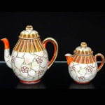 Um(01) bule para chá e uma(01) leiteira em porcelana japonesa, bojo oval assente sobre base circular, decoração policromada e em relevo composta por reservas florais estilizadas sobre fundo perolizado, faixa superior ornamentada por filetes verticais em laranja, azul, verde, branco e dourado, tampa abaulada encimada por puxador em dupla folhagem, bico vertedor curvilíneo, alça lateral curvada. Marca ao fundo JH acompanhada de caracteres japoneses. Medidas: Alt.: 17 x Comp.: 17,5 x Larg.: 11,5 cm. Alt.: 12,5 x Comp.: 12 x Larg.: 8,5 cm.