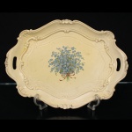 Bandeja de serviço em material plástico moldado, marca Stil, manufatura italiana, identificada no reverso com inscrição Florence Italy e etiqueta original do fabricante. Corpo de formato recortado com alças vazadas integradas, bordas molduradas por elementos de inspiração rocaille em relevo e reserva central decorada por ramalhete de miosótis atado por laço. Reverso com flor-de-lis em relevo. Apresenta sinais de uso, desgastes e marcas do tempo compatíveis com a conservação da peça. Medidas: Comp.: 43 x Larg.: 30,5 cm.