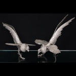 Duas(02) esculturas representando aves em posição de pouso, executadas em metal fundido com acabamento prateado. Corpos modelados com representação de plumagem, asas abertas e patas apoiadas sobre superfície plana. Medidas: Alt.: 27,5 x Comp.: 40,5 x Larg.: 25 cm. ; Medidas: Alt.: 23 x Comp.: 42 x Larg.: 25 cm.