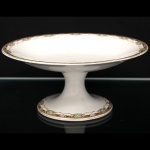 Fruteira de pedestal em porcelana, com prato circular raso apoiada sobre haste central e base circular. Decoração em faixa perimetral policroma composta por elementos fitomórficos, volutas, medalhões e filetes dourados. Peça marcada na base Johnson Bros. England. Inglaterra, século XX. Apresenta sinais de uso compatíveis com o tempo. Medidas: Alt.: 11 x Diam.: 24,5 cm.