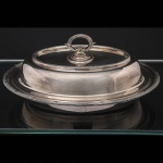 FRACALANZA. Elegante terrina com tampa confeccionada em metal espessurado à prata, para serviço de  vegetais, formato oval de centro rebaixado, borda ornamentada com fileira de frisos relevados, intercalado por fitas, padrão império francês, obtidos através da técnica do cinzelado, ornato replicado na tampa, com puxador removível tornando a tampa um segunda travessa. Marca da manufatura no verso. Med.: Alt. 7 x Comp. 27 x Larg. 21 cm. Apresenta marcas de uso.