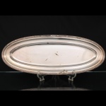 FRACALANZA. Elegante travessa oblonga confeccionada em metal espessurado à prata, formato circular, centro ligeiramente rebaixado, borda relevada com fileira de frisos intercaladas por fitas cruzadas, ornato obtido através da técnica do cinzelado. Marca da manufatura no verso. Apresenta marcas de uso e desgastes. Medida: Comp.: 61 x Larg.: 24 cm.