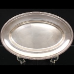 FRACALANZA. Elegante travessa oval confeccionada em metal espessurado à prata, formato circular, centro ligeiramente rebaixado, borda relevada com fileira de frisos intercaladas por fitas cruzadas, ornato obtido através da técnica do cinzelado. Marca da manufatura no verso. Apresenta marcas de uso. Medida: Comp.: x Larg.:   cm. Apresenta marcas de uso.