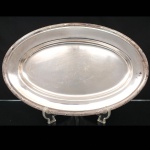 FRACALANZA. Elegante travessa oval confeccionada em metal espessurado à prata, formato circular, centro ligeiramente rebaixado, borda relevada com fileira de frisos intercaladas por fitas cruzadas, ornato obtido através da técnica do cinzelado. Marca da manufatura no verso. Apresenta marcas de uso. Medida: Comp.: x Larg.:   cm. Apresenta marcas de uso.