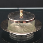 Manteigueira com tampa, composta por recipiente circular em vidro moldado e suporte em metal prateado. Corpo cilíndrico com fundo decorado por motivo radial moldado em relevo. Tampa circular em metal prateado com pega central torneada. Medidas: Alt.: 7 x Diam.: 12 cm.