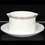 Porcelana Renner, Porto Alegre - RS(Fundada em 1937). Molheira em porcelana nacional da década de 1960/70; esmaltação na cor branca, formato cilíndrico fixada sobre presentoir circular, borda ornada por cercadura em meandro contínuo, de tonalidade cinza, e sucessão de quadrados duplos em douração aplicada, dotada de vertedouros opostos revirados para fora. Marca da manufatura no verso. Medidas: Alt.: 5,5  x Diam.: 21,5 cm.
