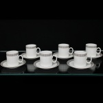 Porcelana Renner, Porto Alegre - RS(Fundada em 1937). Conjunto de xícaras para café em porcelana nacional da década de 1960/70; esmaltação na cor branca, bojo cilíndrico e alça lateral oval aplicada, borda ornada por cercadura em meandro contínuo, de tonalidade cinza, e sucessão de quadrados duplos em douração aplicada, padrão replicado no pires. Marca da manufatura no verso. Composto de seis(06) unidades. Medida: Alt.: 5,5 x Comp.: 7 x Larg.: 5 cm.