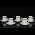 Porcelana Renner, Porto Alegre - RS(Fundada em 1937). Conjunto de xícaras para café em porcelana nacional da década de 1960/70; esmaltação na cor branca, bojo cilíndrico e alça lateral oval aplicada, borda ornada por cercadura em meandro contínuo, de tonalidade cinza, e sucessão de quadrados duplos em douração aplicada, padrão replicado no pires. Marca da manufatura no verso. Composto de seis(06) unidades. Medida: Alt.: 5,5 x Comp.: 7 x Larg.: 5 cm. Uma apresenta um bicado na borda.