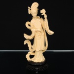 Escultura representando figura feminina oriental, executada em resina moldada de tonalidade marfim. Produção do século XX, de inspiração orientalista, apresentando figura em pé com vestes de tradição chinesa, segurando ramo floral e medalhão, com adorno capilar composto por flores e cacho de uvas. Composição em formato vertical, assentada sobre base circular vazada em material sintético de coloração escura. Decoração composta por panejamento, motivos florais, elementos fitomorfos e atributos simbólicos orientais. Medidas: Alt.: x Comp.: x Larg.: