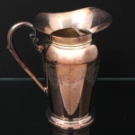 EBERLE. Elegante jarra confeccionada em metal espessurado à prata, bojo balaústre ricamente lavrada com elementos vegetalistas, assente sobre base circular repuxada em degraus, asa lateral em volutas, boda ondulada e revirada para fora, interior dotado de barra retentora de gelo. Marca da manufatura ao fundo. Med.: Alt. 22 x Comp. 20 x Larg. 13 cm. Apresenta marcas de uso e necessita polimento. 