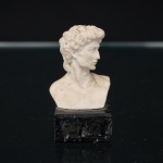 Busto decorativo representando David, personagem da iconografia renascentista, executado em material sintético de tonalidade marfim, execução de inspiração renascentista italiana, do original de Michelangelo, em formato escultórico de meio-busto, assentado sobre base prismática em mármore preto. Medidas: Alt.: 7 x Comp.: 3,5 x Larg.: 3 cm.