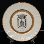 Prato comemorativo de parede, executado em porcelana, destinado à ornamentação e exposição mural. Produção do século XX, de caráter institucional e comemorativo, em formato circular com aba ondulada. Decoração composta por brasão heráldico da cidade de Soria, inscrição Soria Pura Cabeza de Extremadura, cercadura interna com motivos florais gravados em douração e borda com frisos dourados. Marcado no verso Platería Lugones  Oro, Plata, Platino  Oviedo Medida: Diam.: 25 cm.