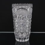 Vaso em cristal incolor moldado e lapidado, de formato cilíndrico levemente afunilado, decorado por reservas arqueadas, reticulado geométrico, losangos, prismas e motivos estrelados. Borda recortada e fundo trabalhado em composição geométrica facetada. Peça destinada à ornamentação floral. Medidas: Alt.: 21,5 x Diam.: 11,5 cm.