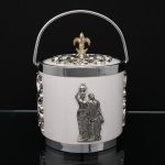 Balde para gelo em material sintético de tonalidade marfim, com tampa e alça móvel em metal cromado. Corpo cilíndrico ornamentado com aplicações em relevo representando figuras femininas em trajes clássicos e elementos vazados de volutas. Tampa com galeria vazada e pegador em forma de flor-de-lis. Medidas: Alt.: 21 x Diam.: 17,5 cm.