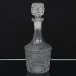 Garrafa licoreira em cristal moldado e lapidado, de corpo cilíndrico ornamentado por frisos verticais, reservas circulares e faixa decorativa em relevo geométrico. Gargalo alongado e afunilado, acompanhado de tampa com lapidação em malha de losangos. Medidas: Alt.: 28,5 x Diam.: 11,5 cm.