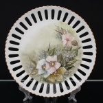 Prato decorativo em porcelana vazada, borda recortada com filete dourado, ornado por pintura floral policromada executada à mão, representando flores e folhagens. Medida: Diam.: 27 cm.